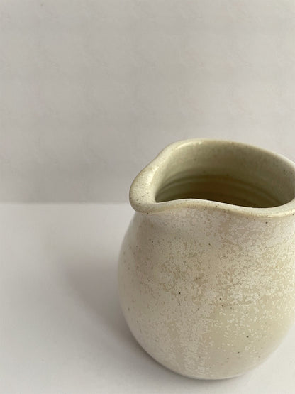 A hand-thrown ceramic milk jug in a beige speckled glaze on a neutral background - close-up view.