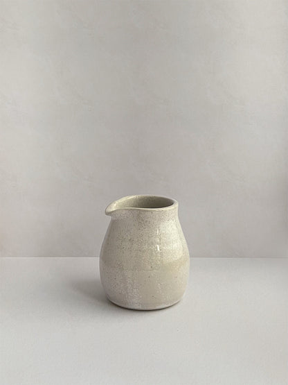 A hand-thrown ceramic milk jug in a beige speckled glaze on a neutral background - left side view.