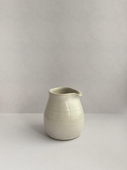 A hand-thrown ceramic milk jug in a beige speckled glaze on a neutral background - right side view.