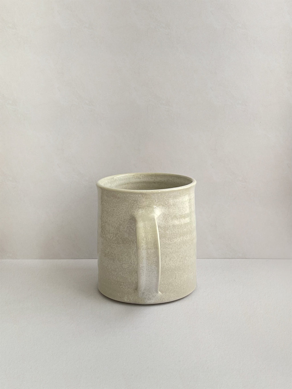 A hand-thrown ceramic mug with a handle, in a beige speckled glaze on a neutral background - back view.
