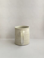 A hand-thrown ceramic mug with a handle, in a beige speckled glaze on a neutral background - back view.