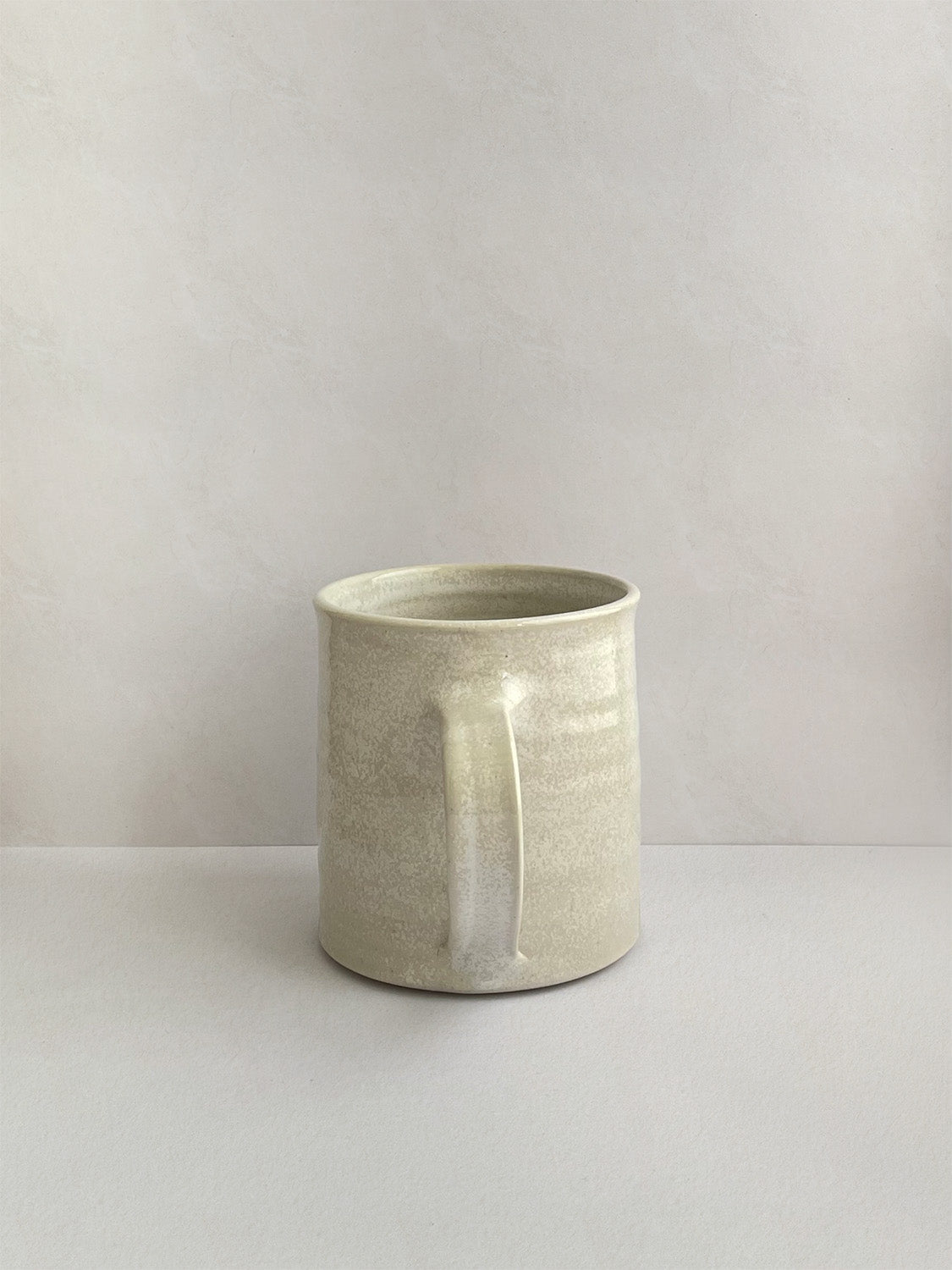 A hand-thrown ceramic mug with a handle, in a beige speckled glaze on a neutral background - back view.