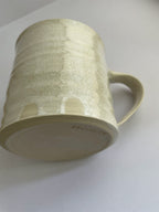 A hand-thrown ceramic mug with a handle, in a beige speckled glaze on a neutral background - detail view.