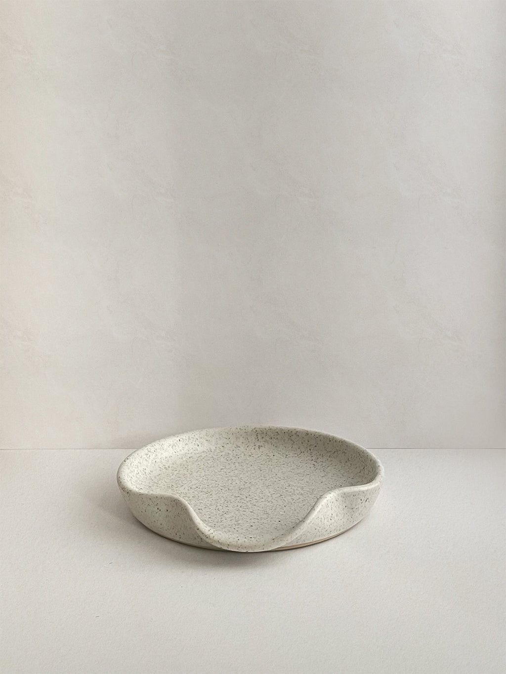 Speckled white handmade ceramic spoon rest on a neutral background - front view.