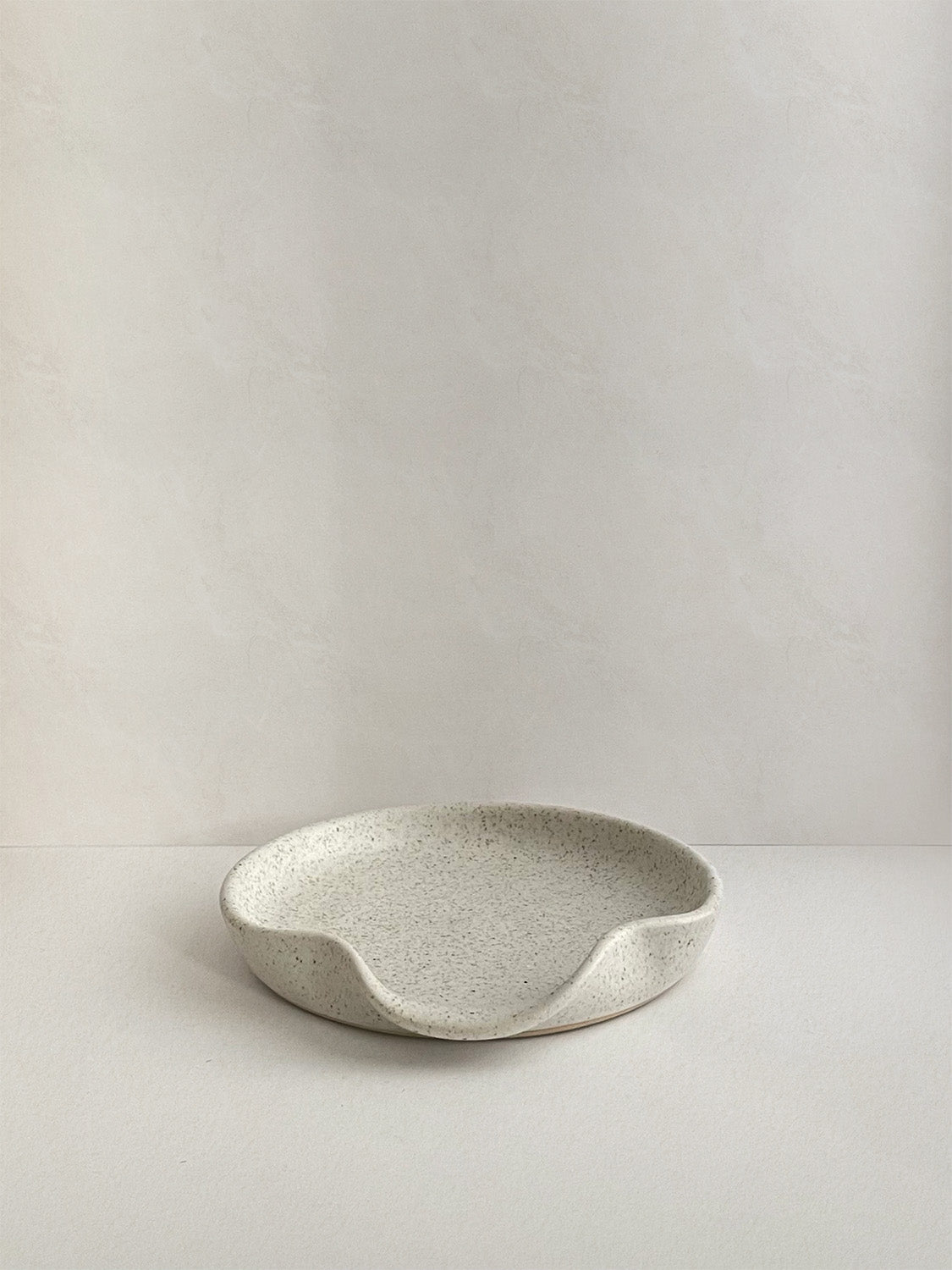Speckled white handmade ceramic spoon rest on a neutral background - front view.