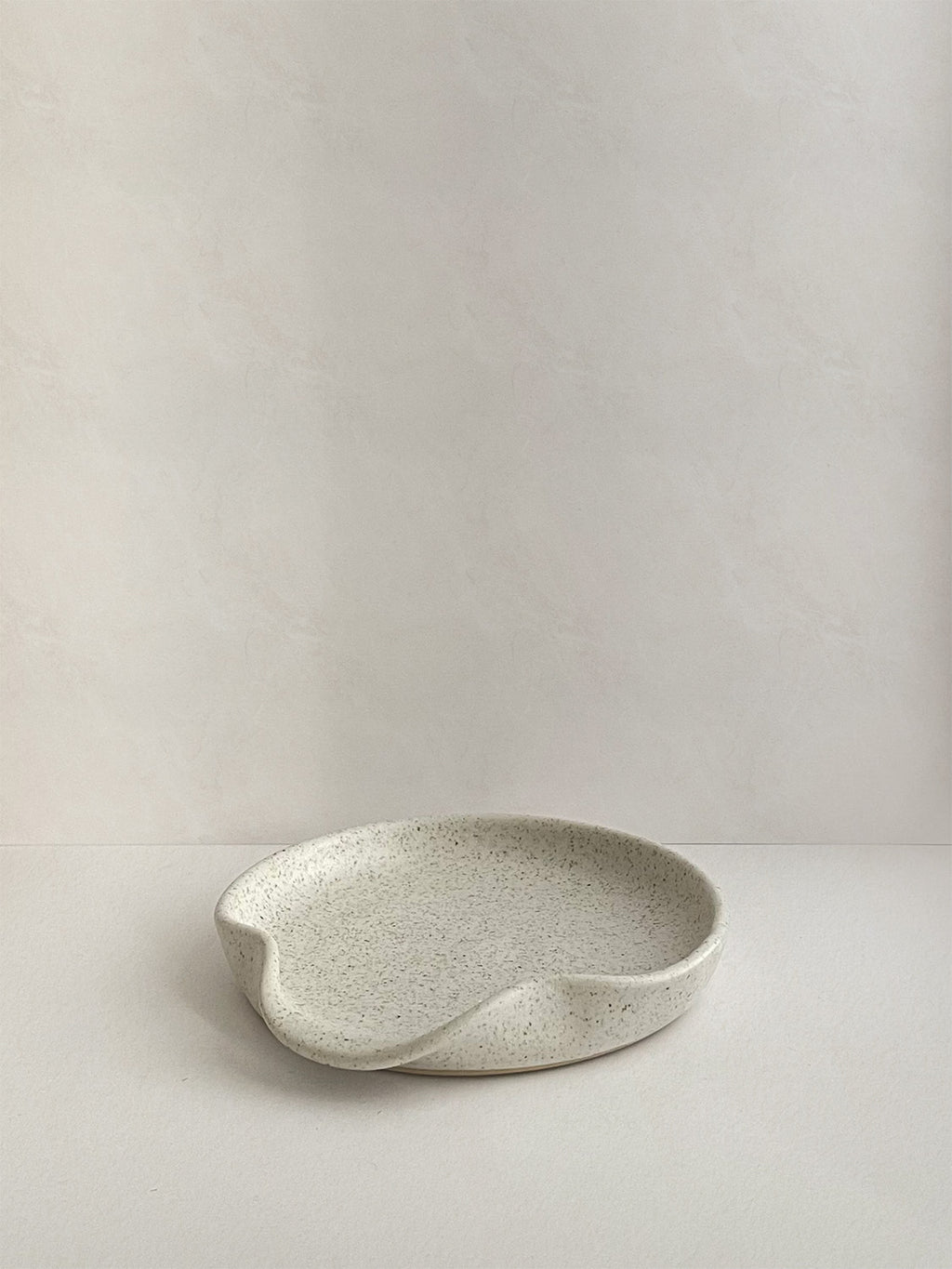 Speckled white handmade ceramic spoon rest on a neutral background - front view.