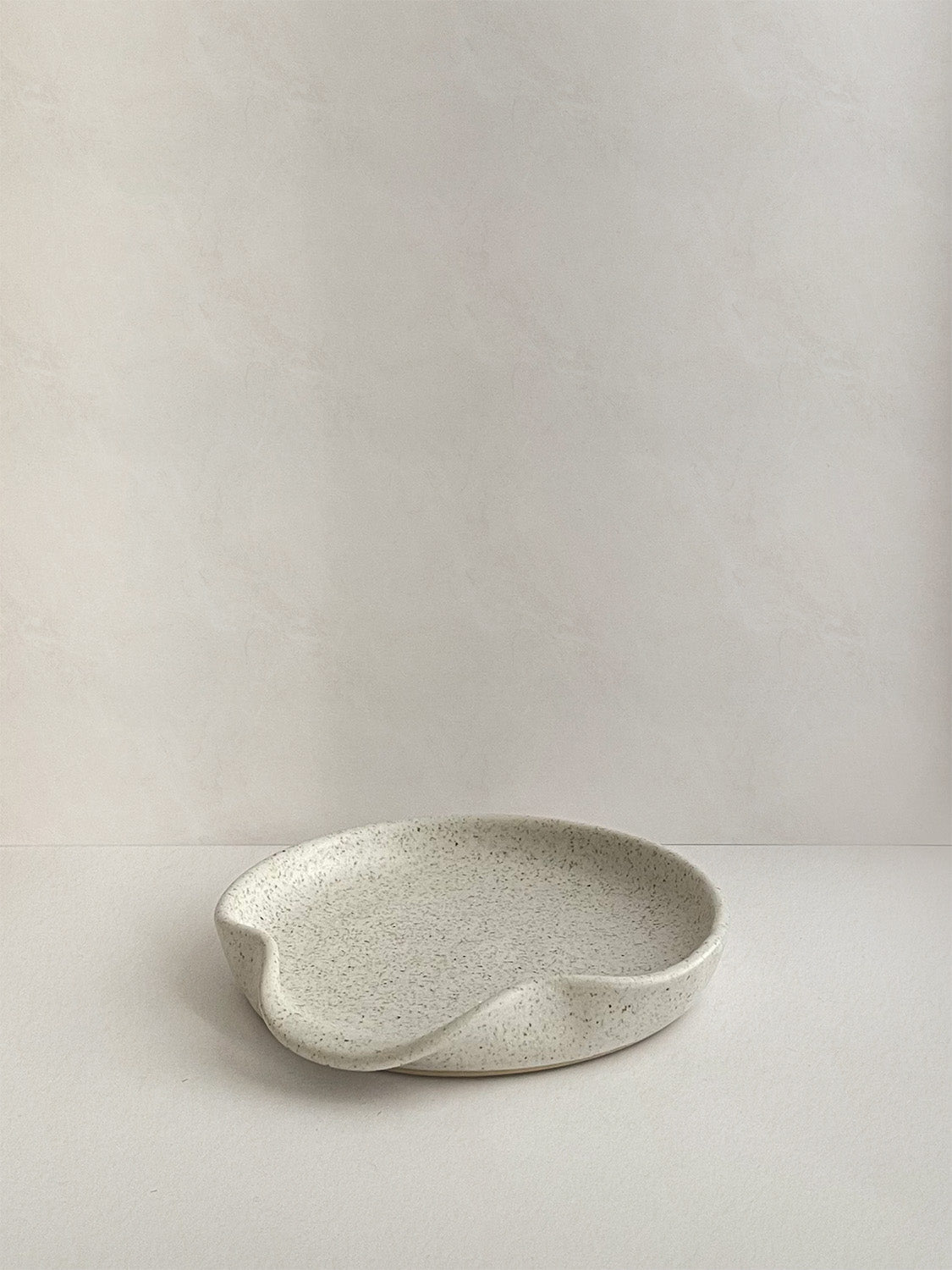 Speckled white handmade ceramic spoon rest on a neutral background - front view.