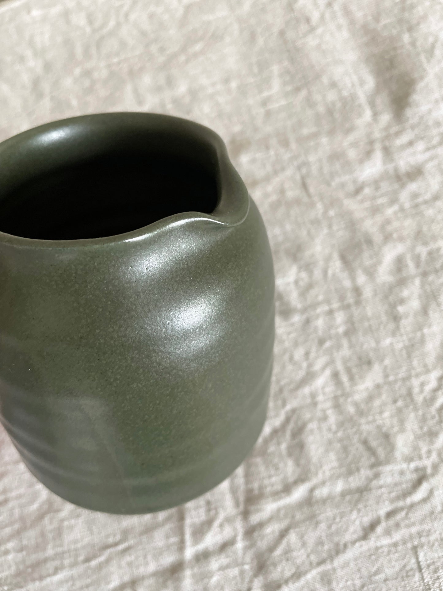 Ceramic jug with handle in deep green glaze on a beige linen background - close-up view.