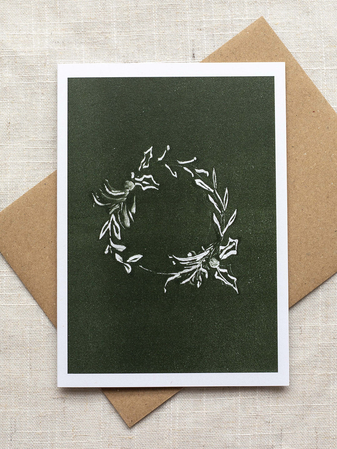Botanical Christmas card with a leafy wreath design on a green background, placed on a linen surface with a Kraft paper envelope.
