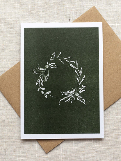 Botanical Christmas card with a leafy wreath design on a green background, placed on a linen surface with a Kraft paper envelope.