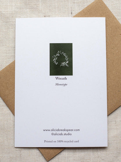 Reverse side of botanical Christmas card with a leafy wreath design on a green background, placed on a linen surface with a Kraft paper envelope.