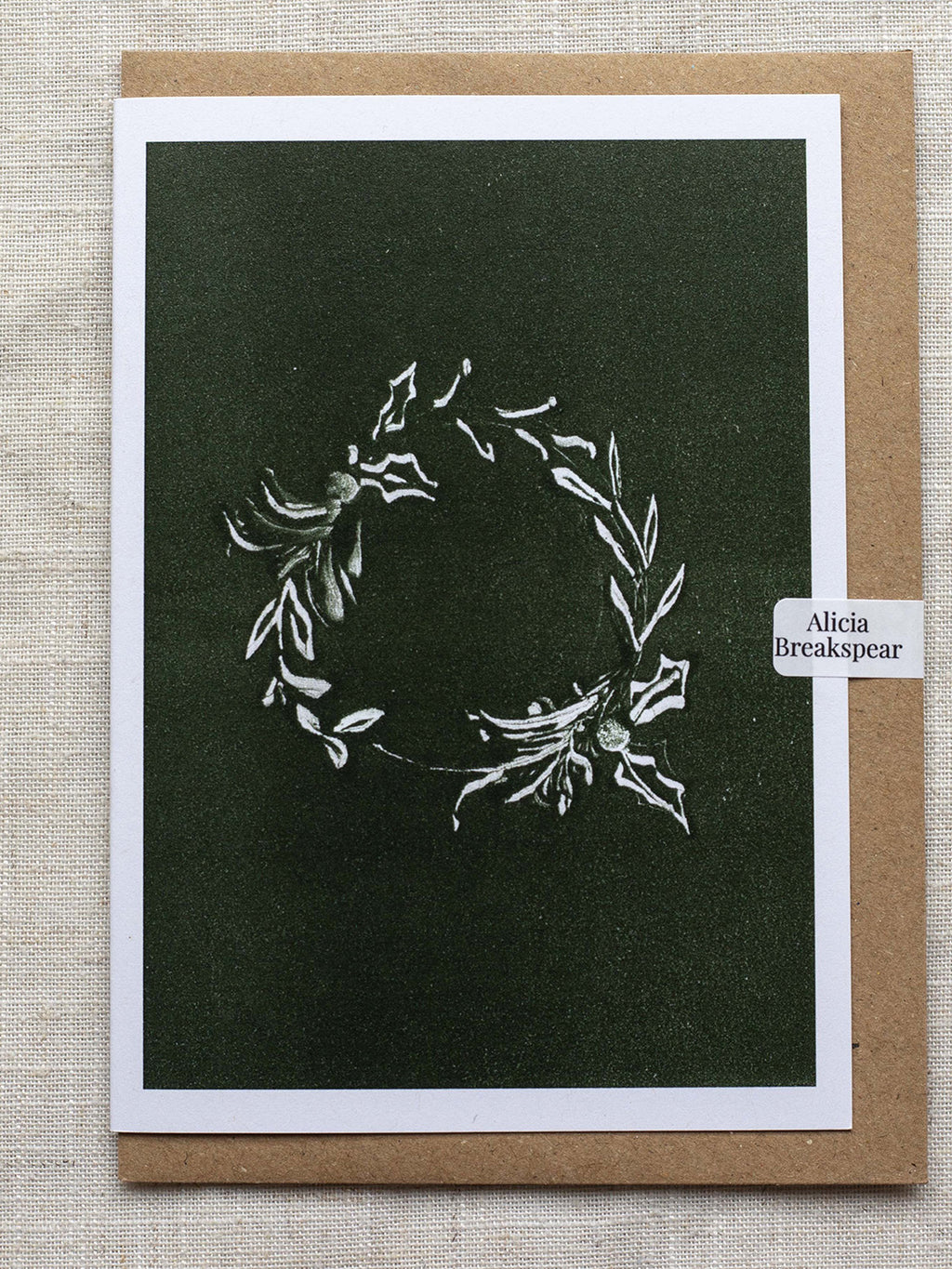 Botanical Christmas card with a leafy wreath design on a green background, with a Kraft paper envelope and 'Alicia Breakspear' sticker seal.