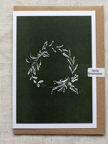 Botanical Christmas card with a leafy wreath design on a green background, with a Kraft paper envelope and 'Alicia Breakspear' sticker seal.