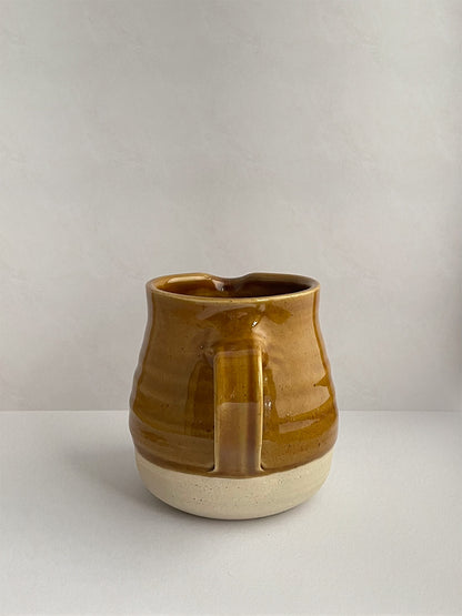 A golden yellow handmade ceramic jug with a curved body and a handle, on a neutral background - back view.
